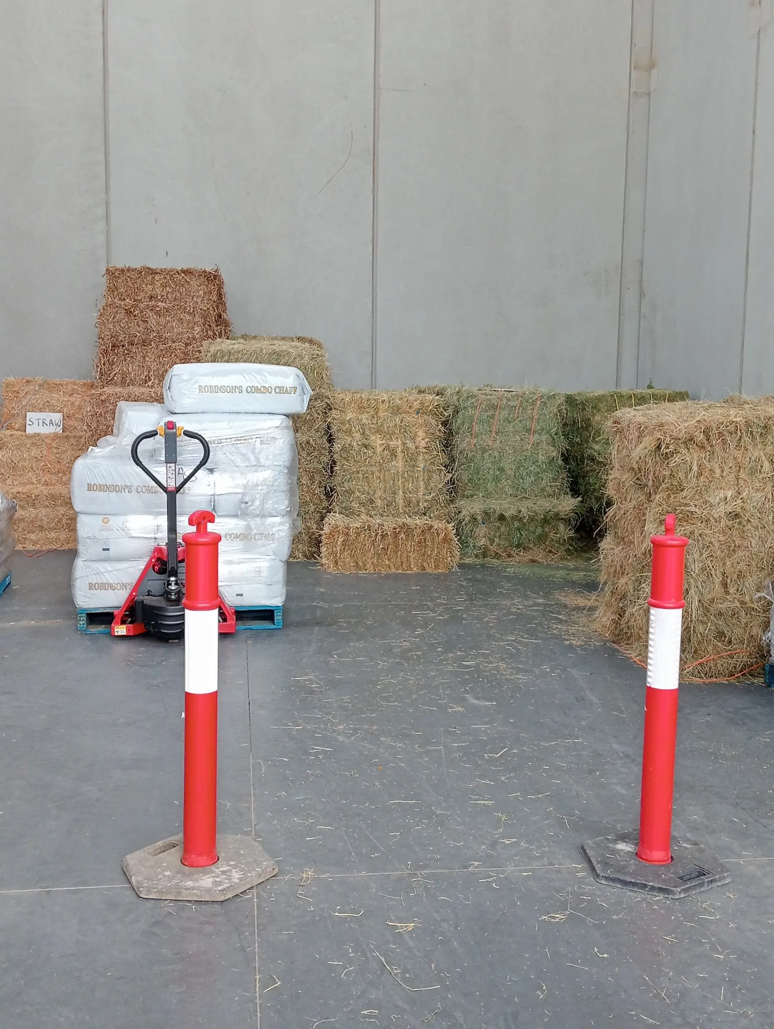 Storage bay with stacked hay and straw bales, red pallet jack, and safety bollards on concrete floor